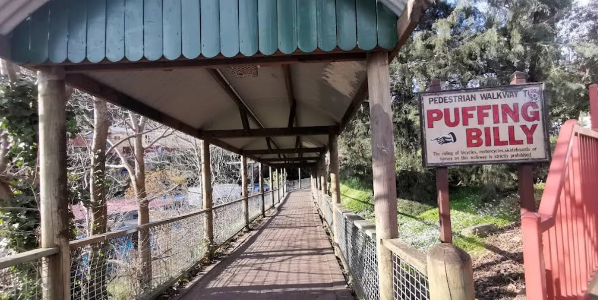 Covered wooden pedestrian walkway ramp with wire fencing and a blue directional sign pointing toward Puffing Billy.