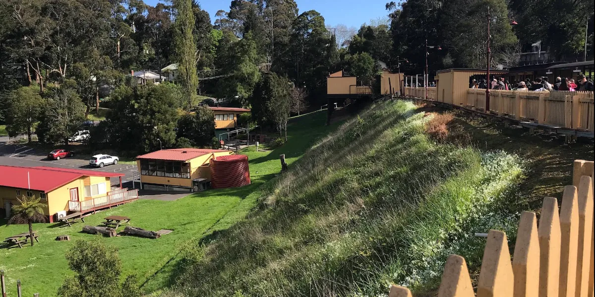 Elevated timber railway station platform with passengers above a sloping lawn with yellow buildings and red roofs.