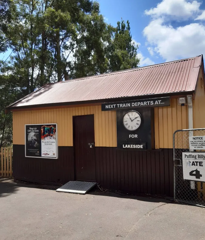 Yellow timber station shed with a next train departure clock to Lakeside beside an open wire security gate labeled Gate 4.