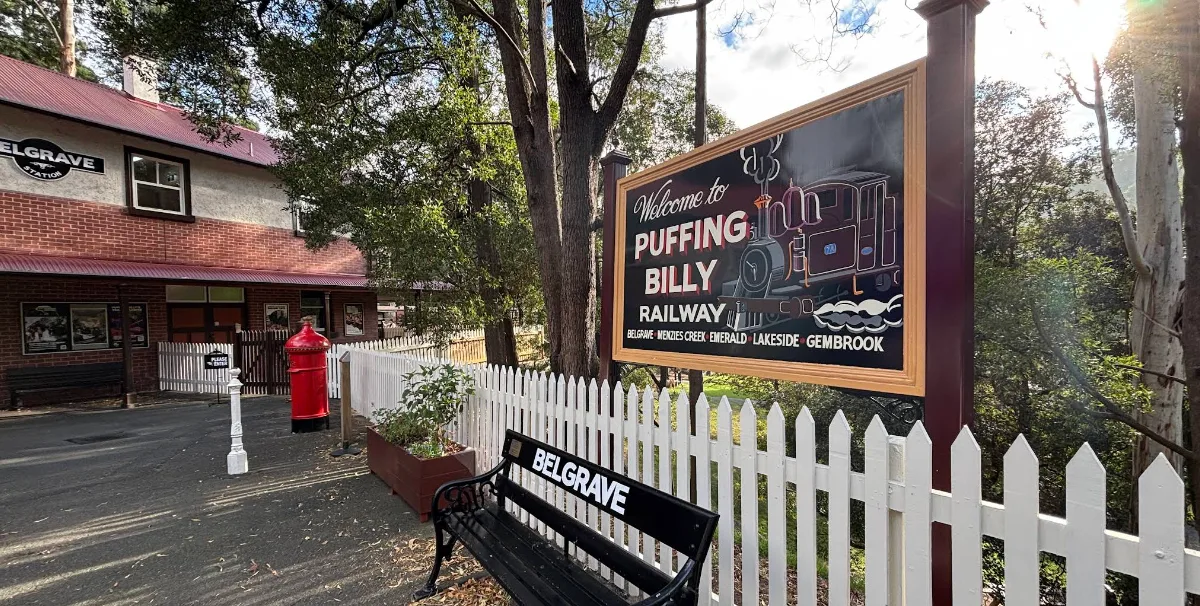 Wooden Puffing Billy Railway welcome sign behind a white picket fence with a red post box and a green Belgrave station bench.